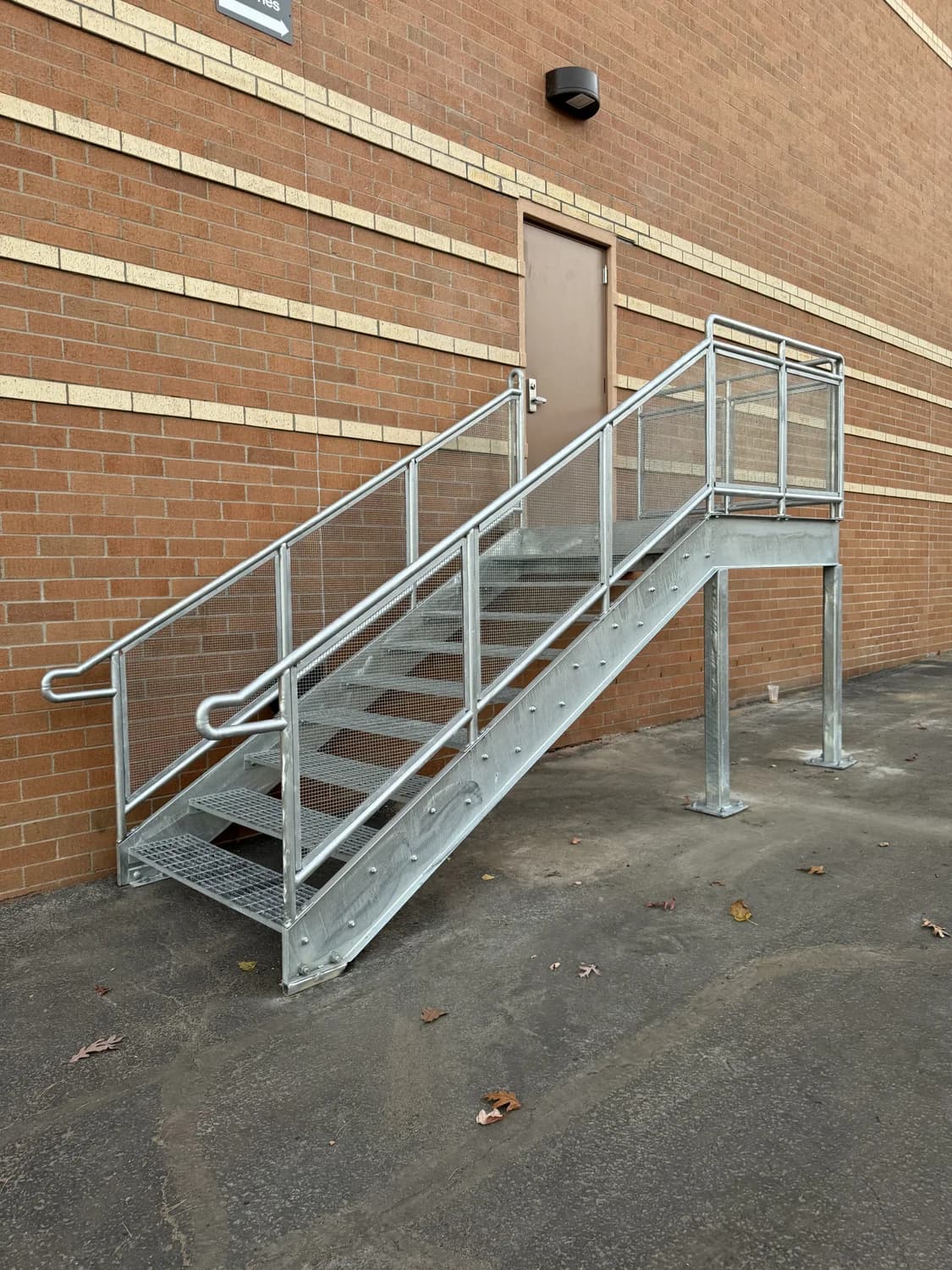 Exterior galvanized steel staircase with grated treads and mesh infill railings beside a brick building
