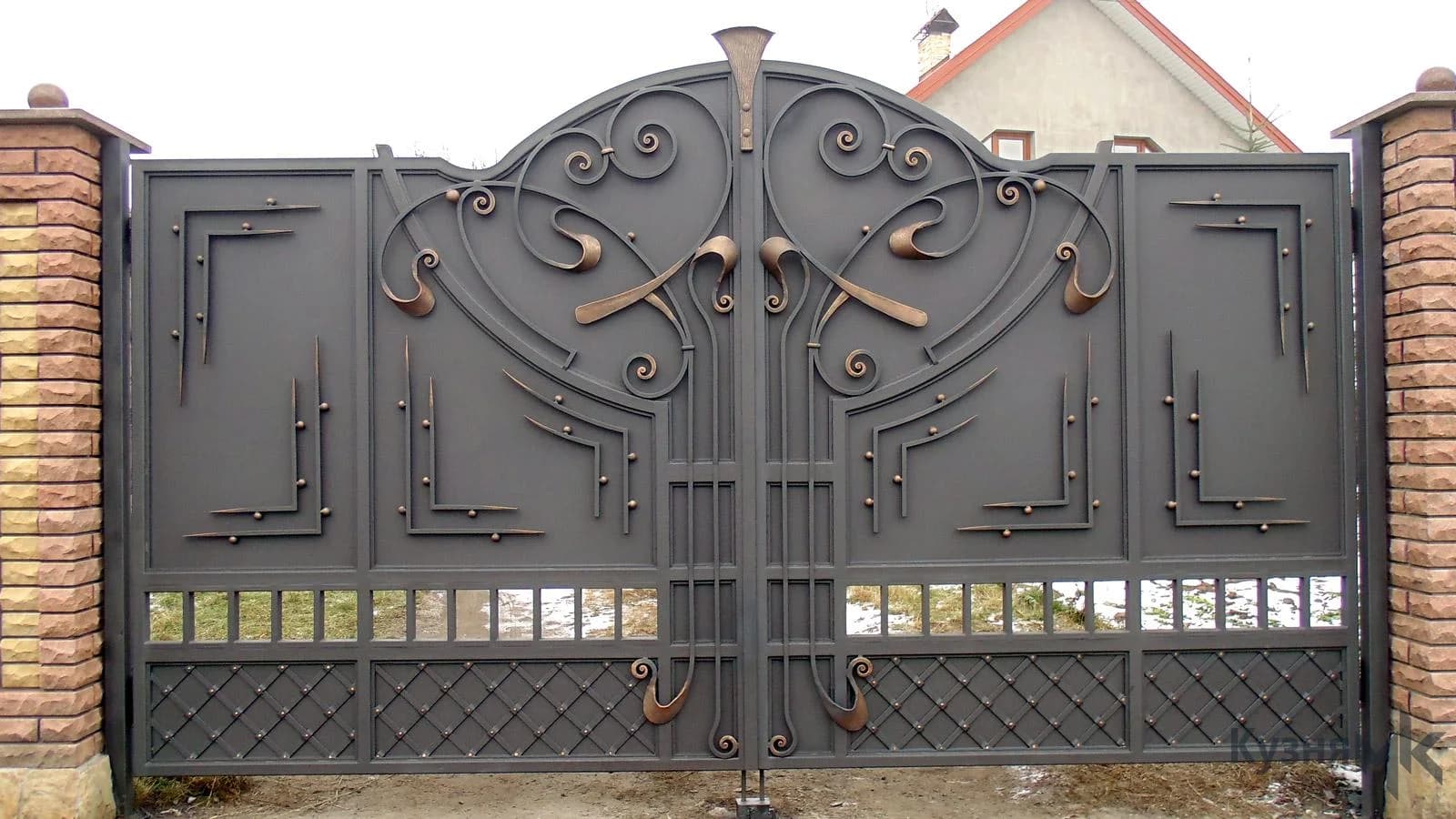 Large decorative metal gate with sculptural scrolls and bronze-colored accents between brick pillars